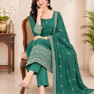 May include: A deep emerald green three-piece ensemble consisting of a long-sleeved kurta with a square neckline, matching straight-leg trousers, and a coordinating dupatta, all featuring intricate silver embroidery and the text Bageshwarexports.