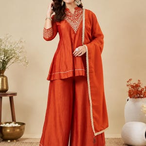 May include: A rust-orange three-piece ensemble consisting of a peplum-style tunic with intricate white embroidery around the neckline and sleeve cuffs, wide-leg trousers with a subtle hemline detail, and a matching dupatta with a delicate lace border.