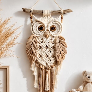 Puede incluir: Un tapiz de macramé con forma de búho hecho de cuerda color crema y tostado, colgado de un trozo de madera flotante. El búho presenta grandes ojos circulares de color marrón oscuro, un pico puntiagudo, alas texturizadas y flecos.