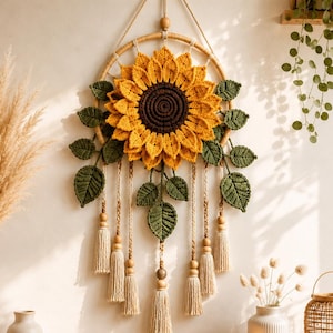 May include: A wall hanging featuring a large crocheted sunflower with a dark brown center and bright yellow petals, mounted on a circular frame. Green crocheted leaves and several hanging beige yarn tassels with wooden beads are attached below.