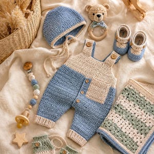 May include: A crochet baby set featuring a blue and cream overall with wooden buttons, a matching blue bonnet, blue and cream booties, green and cream mittens, a bear-shaped wooden rattle, a pacifier clip with wooden beads, and a striped blanket.