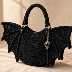 Könnte beinhalten: Eine schwarze gehäkelte Handtasche in Form von Fledermausflügeln mit einem geschwungenen Tragegriff. Die Tasche hat an einer Seite einen goldenen Metallring, an dem ein dekorativer goldener Anhänger mit einem schwarzen ovalen Stein hängt.