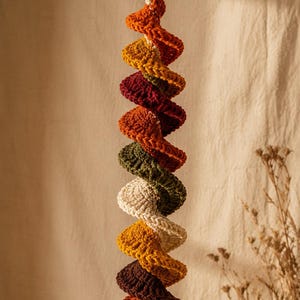 May include: A hanging spiral decoration made of crocheted yarn in a vertical sequence of autumn-inspired colors including burnt orange, mustard yellow, deep burgundy, olive green, and cream, suspended from a smooth, circular wooden ring.
