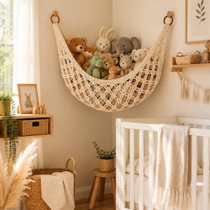 Op de afbeelding: Een crèmekleurige macramé speelgoedhangmat opgehangen in een hoek met twee houten ringen, gevuld met diverse knuffeldieren waaronder een bruine beer, een wit konijn, een grijze olifant en een groene dinosaurus, als opbergoplossing.