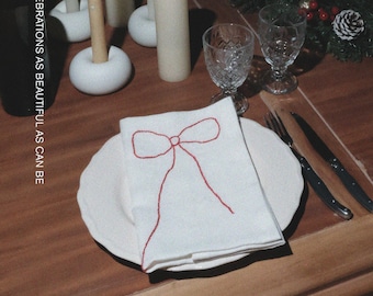 Puede incluir: Una mesa de comedor de madera con platos blancos, vasos transparentes y servilletas blancas con un diseño de lazo bordado en rojo. El texto MŌOBI y MAKE YOUR CHRISTMAS CELEBRATIONS AS BEAUTIFUL AS CAN BE son visibles junto a velas y una corona.