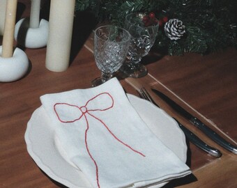 Puede incluir: Una mesa puesta con platos blancos y servilletas bordadas con un lazo rojo. La mesa incluye candelabros blancos con velas, copas de cristal y una corona de pino. El texto dice MŌOBI y MAKE YOUR CHRISTMAS CELEBRATIONS AS BEAUTIFUL AS CAN BE.