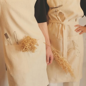 May include: Two cream-colored linen aprons featuring black embroidered line art. One apron displays a whisk and knife, while the other features a dragonfly and a flower. Both aprons have large front pockets and waist ties, worn over black shirts.