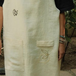 May include: A cream-coloured apron featuring embroidered details including a black insect, a red ladybird, and a yellow leaf outline. The apron includes two front pockets, one of which has a small flap, and is worn over a black short-sleeved shirt.