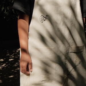 May include: An off-white apron featuring embroidered details including a black moth, a small red ladybug, and a floral stem. The apron has a square neckline with shoulder straps and a large front pocket, crafted from a plain woven fabric.