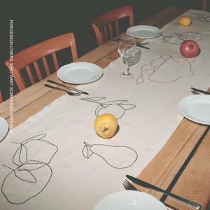 May include: A long, light-colored table runner featuring black line-art illustrations of various fruits, including pears and citrus. The runner is laid across a wooden dining table set with white plates, silverware, a wine glass, and fresh fruit.