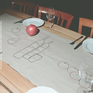 Può includere: Un runner da tavola in lino beige chiaro con ricami a linee nere e rosse raffiguranti bottiglie, frutta e bicchieri. Il testo MŌOBI e FOR DESIGN LOVERS, WHO WANT SOMETHING DIFFERENT è stampato sul runner posizionato su un tavolo in legno.