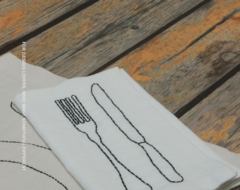 Puede incluir: Una servilleta de tela blanca con un contorno bordado en negro de un tenedor y un cuchillo. El texto MŌOBI y FOR DESIGN LOVERS, WHO WANT SOMETHING DIFFERENT está impreso en la servilleta. Está doblada sobre una superficie de madera.