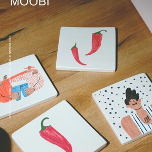 May include: Four square white ceramic coasters featuring hand-painted illustrations. Two coasters display single red chili peppers, one shows a person in a striped shirt with polka dots, and one depicts two figures. The text MŌOBI is visible.