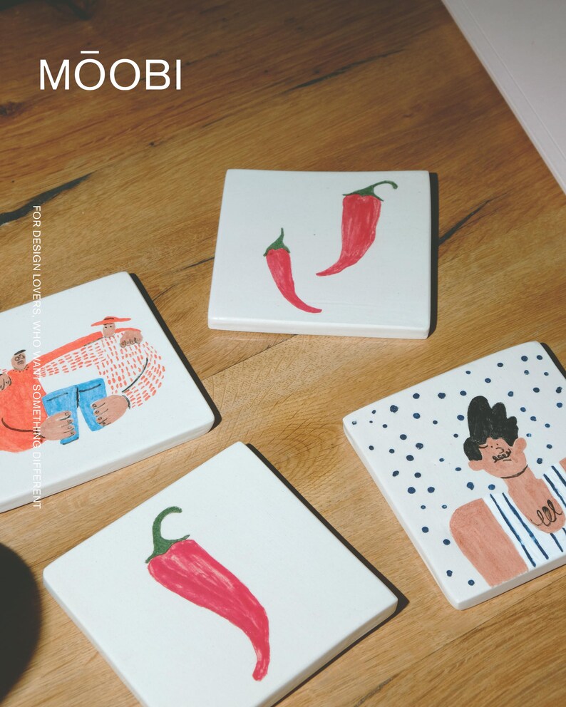 May include: Four square white ceramic coasters featuring hand-painted illustrations. Two coasters display single red chili peppers, one shows a person in a striped shirt with polka dots, and one depicts two figures. The text MŌOBI is visible.