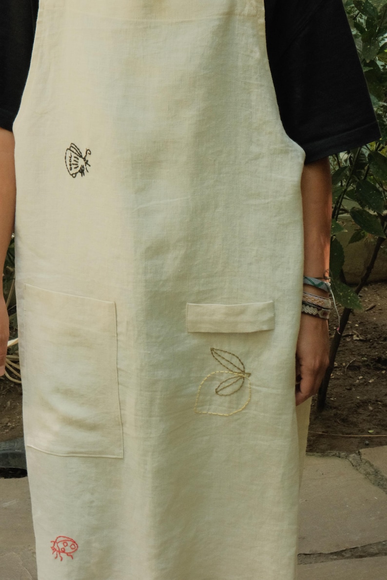 May include: A cream-coloured apron featuring embroidered details including a black insect, a red ladybird, and a yellow leaf outline. The apron includes two front pockets, one of which has a small flap, and is worn over a black short-sleeved shirt.