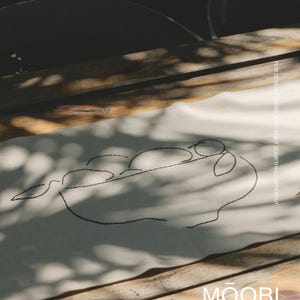 Pu&ograve; includere: Un runner da tavola rettangolare in tessuto color bianco sporco con un ricamo nero che raffigura una ciotola di frutta in stile line art. Il testo "FOR DESIGN LOVERS, WHO WANT SOMETHING DIFFERENT" e il marchio "MOOBI" sono stampati sul tessuto.