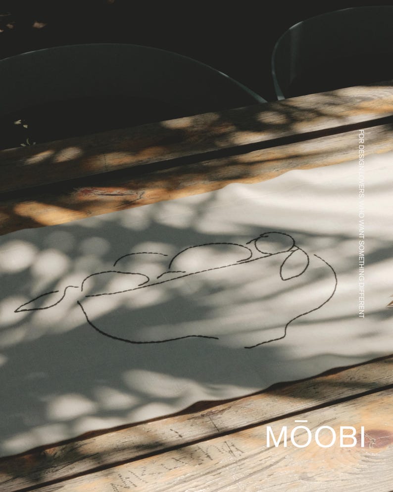 Pu&ograve; includere: Un runner da tavola rettangolare in tessuto color bianco sporco con un ricamo nero che raffigura una ciotola di frutta in stile line art. Il testo "FOR DESIGN LOVERS, WHO WANT SOMETHING DIFFERENT" e il marchio "MOOBI" sono stampati sul tessuto.