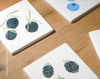 Puede incluir: Tres azulejos de cerámica blancos cuadrados con ilustraciones de frutas pintadas a mano. Un azulejo muestra cinco arándanos azules, otro tres aceitunas oscuras con hojas verdes y el tercero tres moras texturizadas. El nombre de la marca MŌOBI está impreso en la parte inferior.