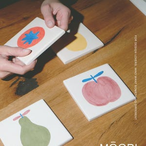 Puede incluir: Cuatro azulejos cuadrados de cer&aacute;mica blanca con ilustraciones de frutas pintadas a mano, incluyendo un tomate rojo con una estrella azul, un c&iacute;rculo amarillo, una manzana rosa con tallo azul y una pera verde con tallo rosa. Texto: FOR DESIGN LOVERS, WHO WANT SOMETHING DIFFERENT y MŌOBI.