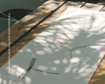 Puede incluir: Un camino de mesa rectangular blanco con delicados bordados de arte lineal de flores, hojas y una libélula. El texto MŌOBI y FOR DESIGN LOVERS, WHO WANT SOMETHING DIFFERENT está impreso en el lado izquierdo del camino de tela.