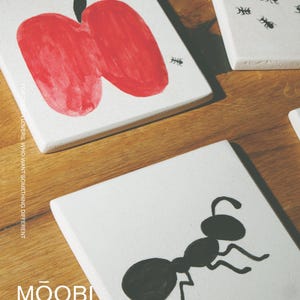 Pu&ograve; includere: Piastrelle quadrate in ceramica bianca con illustrazioni dipinte a mano. Una piastrella mostra una grande mela rossa con un picciolo nero e una piccola formica nera. Un'altra piastrella mostra una grande formica nera. Testo FOR DESIGN LOVERS, WHO WANT SOMETHING DIFFERENT MOOBI.