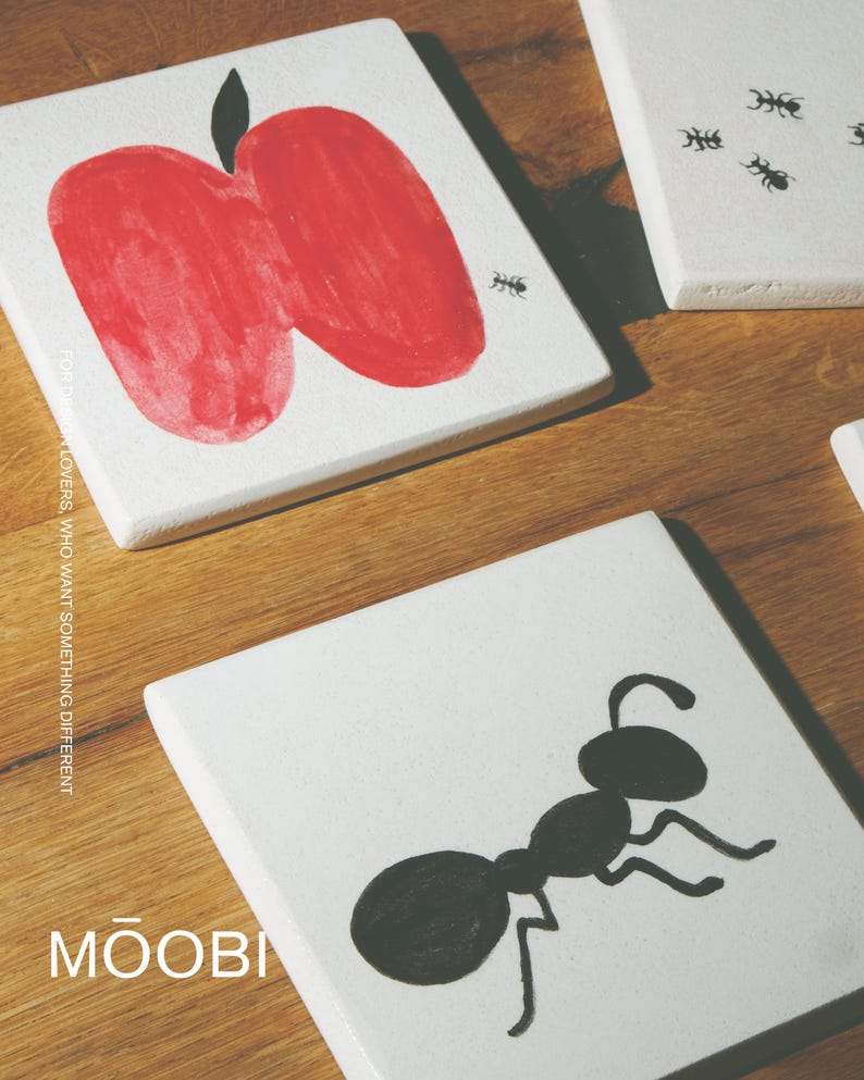 Pu&ograve; includere: Piastrelle quadrate in ceramica bianca con illustrazioni dipinte a mano. Una piastrella mostra una grande mela rossa con un picciolo nero e una piccola formica nera. Un'altra piastrella mostra una grande formica nera. Testo FOR DESIGN LOVERS, WHO WANT SOMETHING DIFFERENT MOOBI.