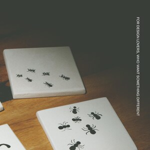 Pu&ograve; includere: Sottobicchieri quadrati bianchi con illustrazioni di formiche nere. Il testo MŌOBI e FOR DESIGN LOVERS, WHO WANT SOMETHING DIFFERENT sono visibili. I sottobicchieri hanno una superficie liscia e sono disposti su una superficie in legno. Ogni sottobicchiere mostra un motivo unico di piccole formiche nere.