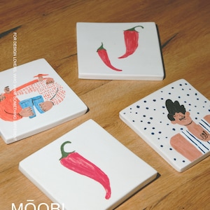 May include: Four square white ceramic coasters featuring hand-painted illustrations. One shows two red chili peppers, another a single red chili pepper, one a person in a striped shirt, and one two figures with the text FOR DESIGN LOVERS, WHO WANT SOMETHING DIFFERENT and MŌOBI.