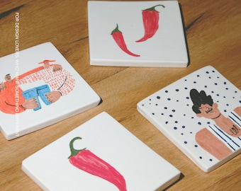 Puede incluir: Cuatro posavasos cuadrados de cerámica blanca con ilustraciones pintadas a mano. Muestran dos chiles rojos, un chile rojo, una persona con camisa de rayas y dos figuras con el texto FOR DESIGN LOVERS, WHO WANT SOMETHING DIFFERENT y MŌOBI.