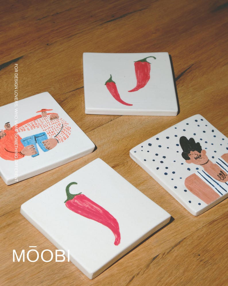 May include: Four square white ceramic coasters featuring hand-painted illustrations. One shows two red chili peppers, another a single red chili pepper, one a person in a striped shirt, and one two figures with the text FOR DESIGN LOVERS, WHO WANT SOMETHING DIFFERENT and MŌOBI.