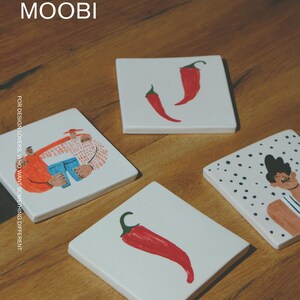 May include: Four square white ceramic coasters featuring hand-painted illustrations. One shows two figures embracing, one features a single red chili pepper, one displays two red chili peppers, and one depicts a person against a black polka dot background.