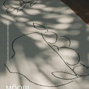 Pu&ograve; includere: Un tessuto bianco con ricami a linee nere che raffigurano un fiore e una ciotola di frutta. Il testo "FOR DESIGN LOVERS, WHO WANT SOMETHING DIFFERENT" &egrave; stampato verticalmente e il marchio "MŌOBI" &egrave; riportato in bianco in basso.