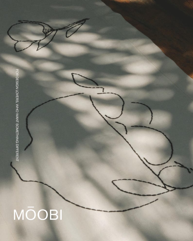Pu&ograve; includere: Un tessuto bianco con ricami a linee nere che raffigurano un fiore e una ciotola di frutta. Il testo "FOR DESIGN LOVERS, WHO WANT SOMETHING DIFFERENT" &egrave; stampato verticalmente e il marchio "MŌOBI" &egrave; riportato in bianco in basso.