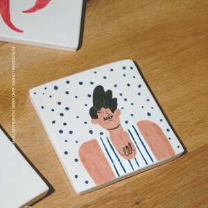 May include: A square white ceramic tile featuring a hand-painted illustration of a person with dark hair and a mustache, wearing a blue and white striped tank top against a background of scattered dark blue dots, with the text MOOBI visible.