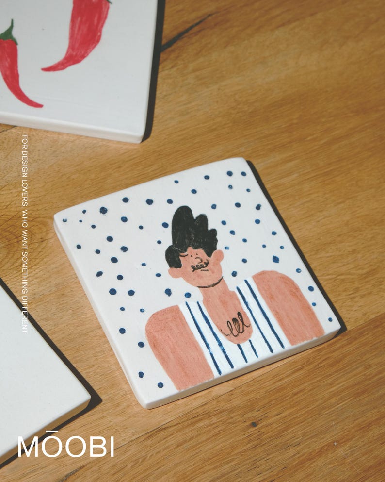 May include: A square white ceramic tile featuring a hand-painted illustration of a person with dark hair and a mustache, wearing a blue and white striped tank top against a background of scattered dark blue dots, with the text MOOBI visible.