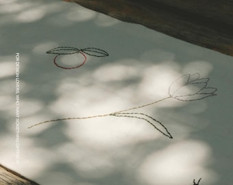 Puede incluir: Un tejido de tela blanca con bordados minimalistas de una flor, una pequeña fruta circular con dos hojas y un pequeño insecto. El texto MOOBI y FOR DESIGN LOVERS, WHO WANT SOMETHING DIFFERENT está impreso en color blanco sobre la tela.