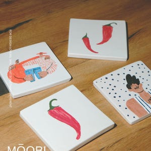 May include: Four square white ceramic coasters featuring hand-drawn illustrations. One shows two figures, one shows two red chili peppers, one shows a single red chili pepper, and one shows a person against a blue polka dot background. Text reads MOOBI.