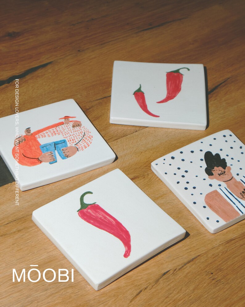 May include: Four square white ceramic coasters featuring hand-drawn illustrations. One shows two figures, one shows two red chili peppers, one shows a single red chili pepper, and one shows a person against a blue polka dot background. Text reads MOOBI.