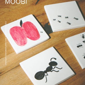 Pu&ograve; includere: Un set di sottobicchieri quadrati bianchi con illustrazioni nere e rosse. Un sottobicchiere mostra una mela rossa con una piccola formica nera, mentre altri mostrano vari disegni di formiche nere. Il testo MŌOBI e FOR DESIGN LOVERS, WHO WANT SOMETHING DIFFERENT &egrave; visibile.