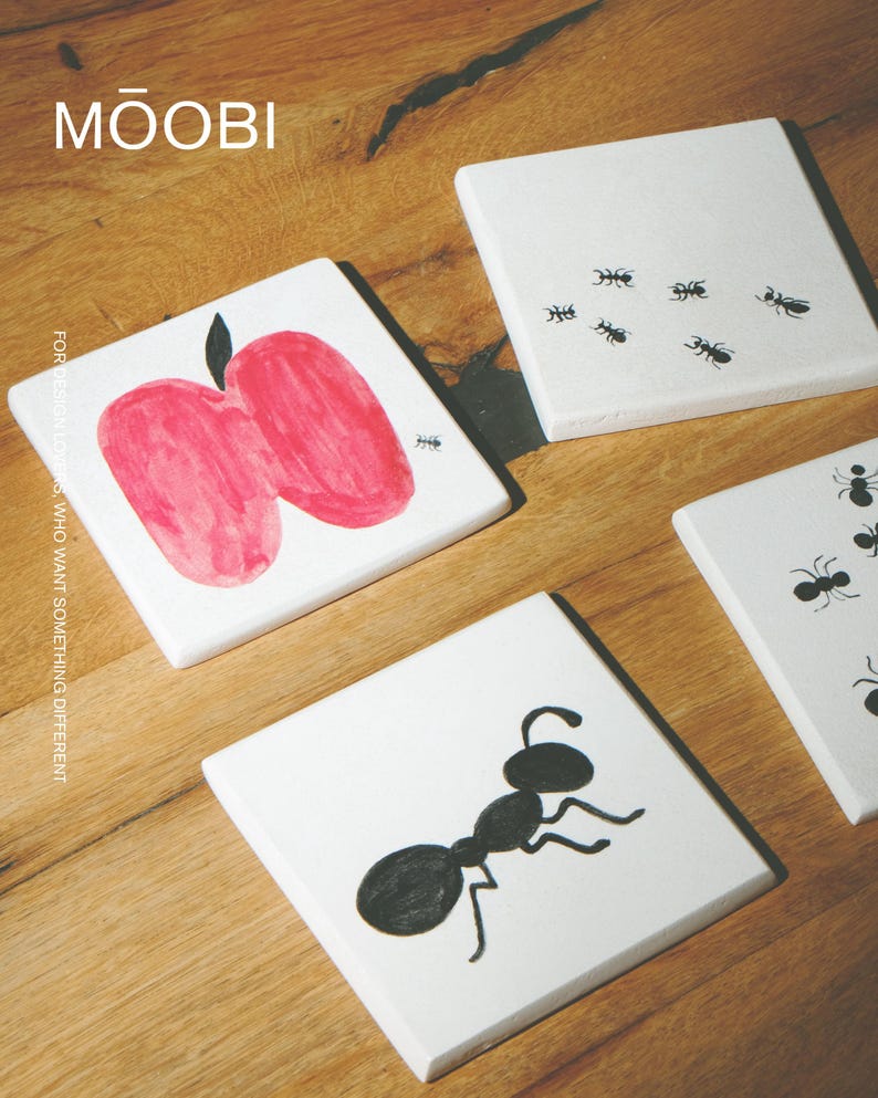 Pu&ograve; includere: Un set di sottobicchieri quadrati bianchi con illustrazioni nere e rosse. Un sottobicchiere mostra una mela rossa con una piccola formica nera, mentre altri mostrano vari disegni di formiche nere. Il testo MŌOBI e FOR DESIGN LOVERS, WHO WANT SOMETHING DIFFERENT &egrave; visibile.