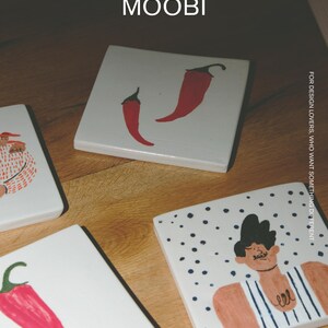 May include: Square white ceramic coasters featuring hand-painted illustrations of red chili peppers and a person in a striped shirt. The text MŌOBI and FOR DESIGN LOVERS, WHO WANT SOMETHING DIFFERENT is printed on the surface of the coasters.