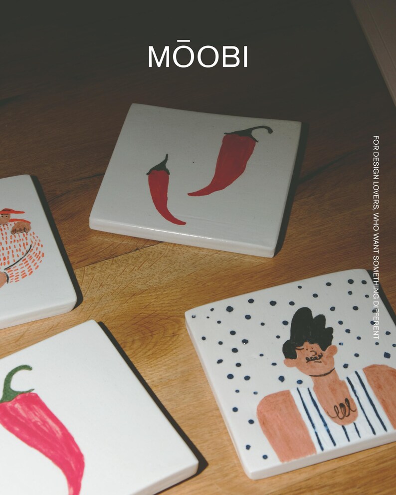 May include: Square white ceramic coasters featuring hand-painted illustrations of red chili peppers and a person in a striped shirt. The text MŌOBI and FOR DESIGN LOVERS, WHO WANT SOMETHING DIFFERENT is printed on the surface of the coasters.