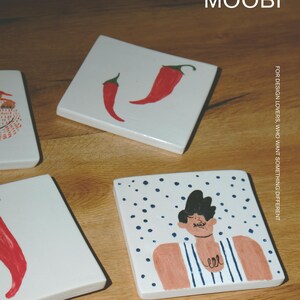 May include: Three square white ceramic tiles featuring hand-painted illustrations. One tile displays two red chili peppers, another shows a partial figure, and the third depicts a person in a blue striped tank top against a blue dotted background. Text reads MŌOBI.