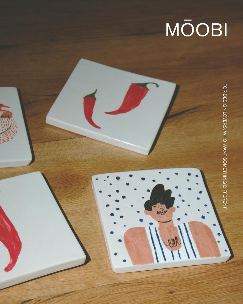 May include: Three square white ceramic tiles featuring hand-painted illustrations. One tile displays two red chili peppers, another shows a partial figure, and the third depicts a person in a blue striped tank top against a blue dotted background. Text reads MŌOBI.