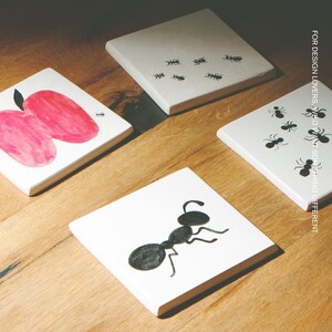 Pu&ograve; includere: Quattro sottobicchieri quadrati in ceramica bianca con illustrazioni a inchiostro nero di formiche e una mela rosa. Un sottobicchiere mostra una grande formica, un altro diverse piccole formiche e uno una mela rosa. Testo: MOOBI e FOR DESIGN LOVERS, WHO WANT SOMETHING DIFFERENT.