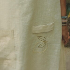 May include: A light cream-colored apron featuring two front pockets with flaps. The fabric is adorned with delicate embroidery, including a stylized green leaf design below one pocket and a small red circular motif near the bottom hem.