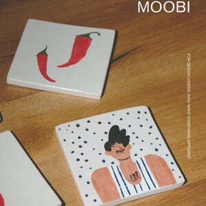 May include: Two square white ceramic coasters. One coaster features two red chili peppers. The other coaster displays a hand-drawn illustration of a person with dark hair wearing a blue and white striped shirt against a background of blue dots. Text reads MŌOBI.