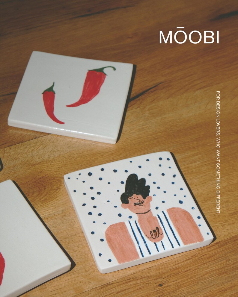 May include: Two square white ceramic coasters. One coaster features two red chili peppers. The other coaster displays a hand-drawn illustration of a person with dark hair wearing a blue and white striped shirt against a background of blue dots. Text reads MŌOBI.