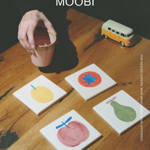 May include: Four square white ceramic coasters featuring hand-painted fruit illustrations including a yellow lemon, a red tomato with a blue center, a green pear, and a red apple. The text MŌOBI appears at the top, with FOR DESIGN LOVERS, WHO WANT SOMETHING DIFFERENT printed vertically on the right side.