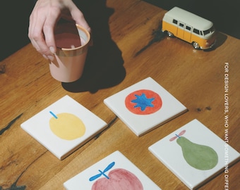 Puede incluir: Cuatro posavasos cuadrados de cerámica blanca con ilustraciones de frutas pintadas a mano, incluyendo un limón amarillo, un tomate rojo con centro azul, una pera verde y una manzana roja. El texto MŌOBI aparece arriba, con FOR DESIGN LOVERS, WHO WANT SOMETHING DIFFERENT impreso verticalmente en el lado derecho.